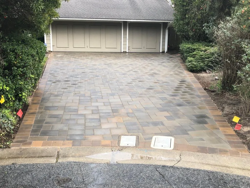 A wet driveway leading to a double garage with closed beige doors, surrounded by lush greenery and bushes in the Bay Area, expertly installed by a paver contractor.
