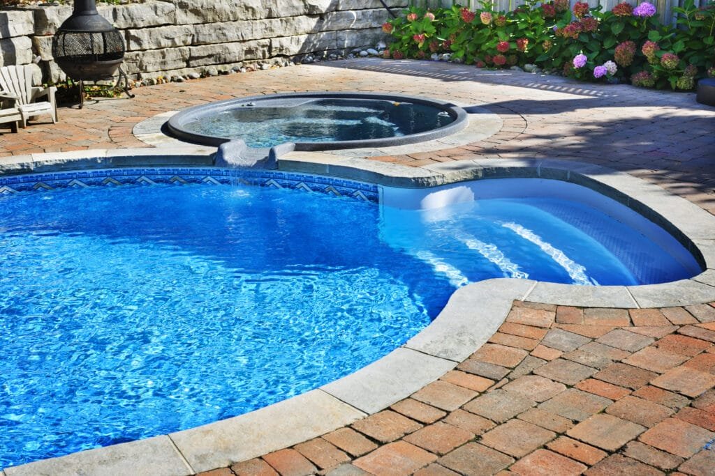 Two adjacent inground pools, one circular hot tub and one larger round pool, surrounded by interlocking paving system and flowering shrubs.