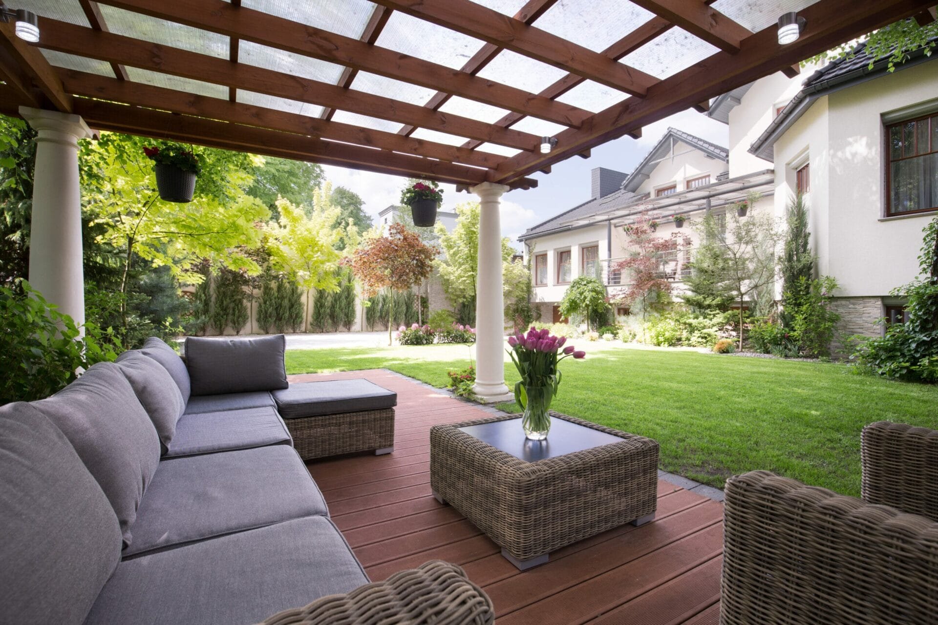 A well-designed patio with a pergola, featuring a cozy seating area, interlocking paving system, hanging plants, and a view of a lush garden and a stately home.