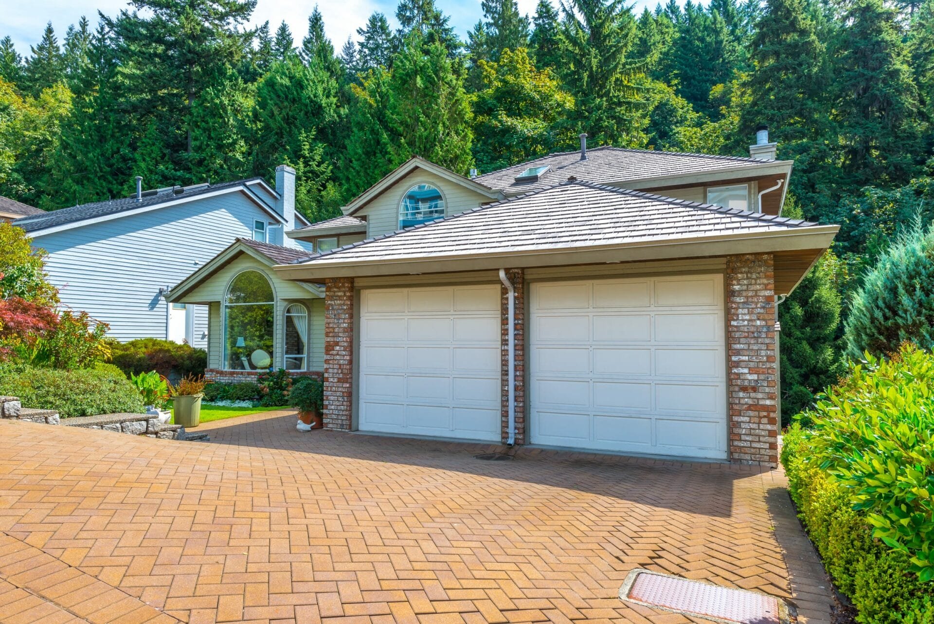 A suburban house with a double garage, white siding, and a shingled roof, surrounded by lush greenery and an interlocking paving system driveway.