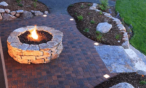 Stone fire pit alight within a curved pathway lined with embedded lights and bordered by landscaping rocks during dusk, enhanced by paving stone installation from an interlocking paving company.